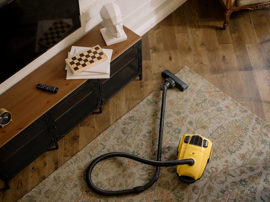 A person is kneeling on a patterned carpet in a living room, using a yellow and black vacuum cleaner to perform deep cleaning. Their hands are holding a white cloth to wipe inside the vacuum's dust co