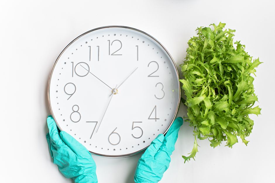 A person wearing turquoise cleaning gloves is holding a large, round wall clock with a white face, black numbers, and black minute and hour hands, showing the time at approximately 12:08. The backgrou
